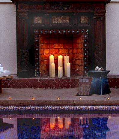 A cozy poolside spa setup with two white lounge chairs, neatly folded towels, a glowing fireplace with candles, and a small table by the side, all reflected in the pool.