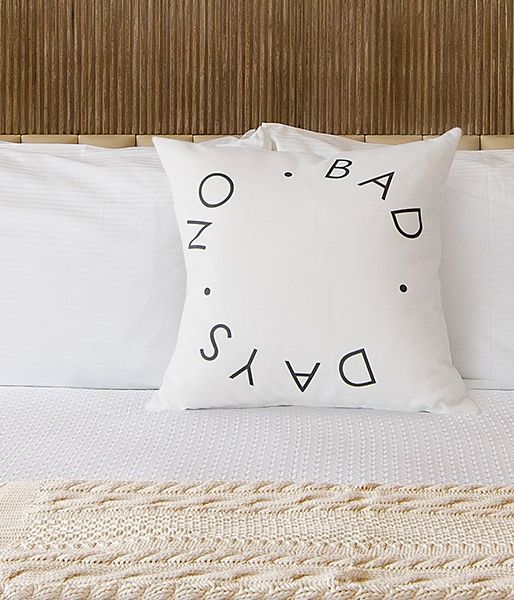 A neatly made bed with white linens, two matching lamps, a textured throw at foot, and a decorative pillow reading &ldquo;DAYS&rdquo; on a wood-paneled headboard.