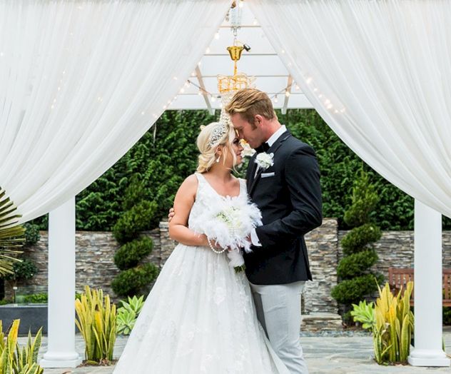 A bride and groom kiss under a white arch in a sunny outdoor wedding setting, bouquet in hand.