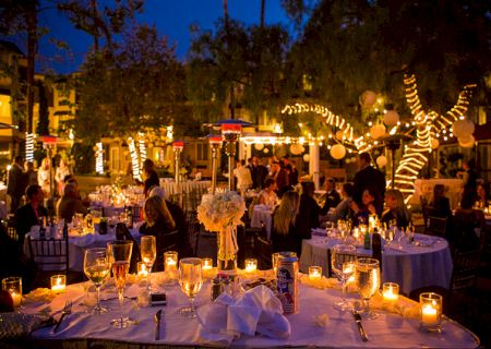 A lively outdoor scene at night with colorful lights, a fountain, palm trees, and people strolling along a warmly lit promenade.
