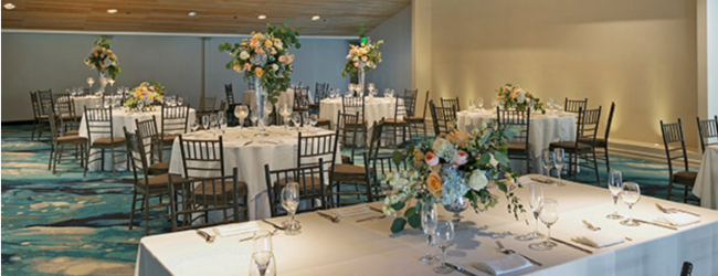 A banquet hall with white-clothed tables, floral centerpieces, elegant place settings, dark wooden chairs, and warm ceiling lights.