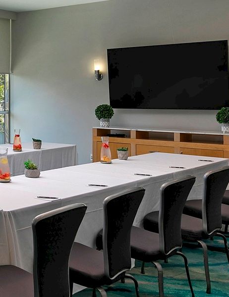 A bright conference room with a U-shaped table set for a meeting, white linens, bottled drinks, snack table by windows, and a mounted TV.