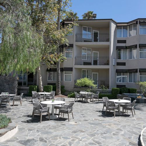 A sunny courtyard with a brick apartment building, stairs to entrances, trees, and parked cars in front, bright and inviting.