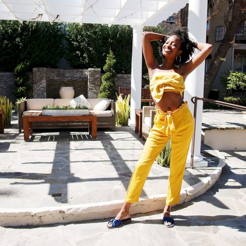 A woman in a yellow two-piece outfit poses confidently near a poolside patio with modern outdoor furniture and greenery, under a white pergola.
