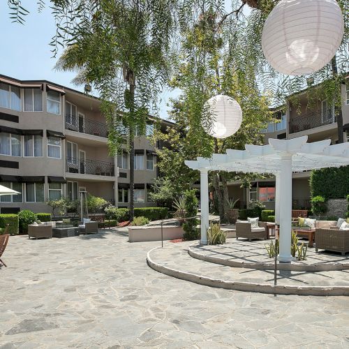 A sunny outdoor patio with wooden tables, chairs, and string lights, surrounded by palm trees and tropical greenery.