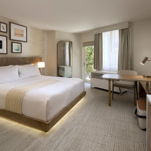 A modern hotel room with a large bed, nightstands, a desk, a chair, a dresser with a TV, neutral tones, artwork above the bed, and a window with drapes.