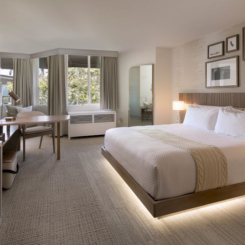 A modern hotel room with a king bed, bedside lamps, framed art above the headboard, a desk and chair, a flat-screen TV, and a seating area by large windows.