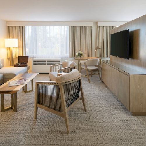 A modern hotel suite with a living area: sofa, armchairs, coffee table, desk, large window, and a wall-mounted TV on a light wood divider.