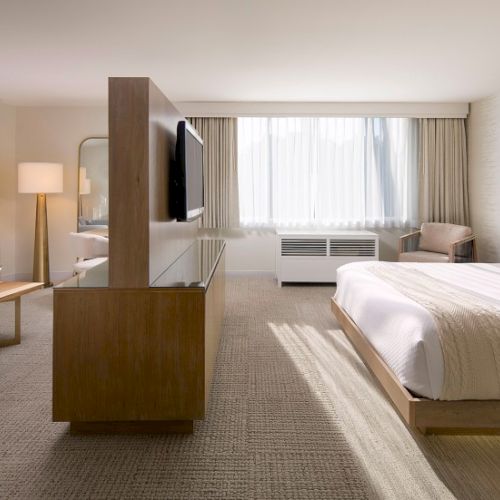 A bright hotel room with two beds, a seating area, and a TV facing a windowed wall, airy and modern.