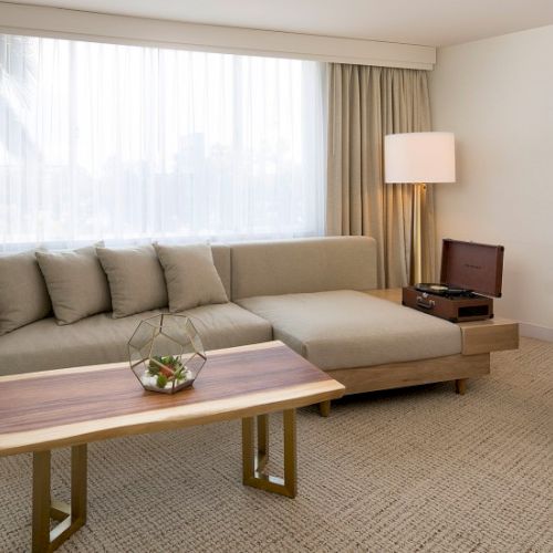 A modern hotel living room with a light gray sectional sofa, wooden coffee table, neutral tones, large window with sheer curtains, and a floor lamp.