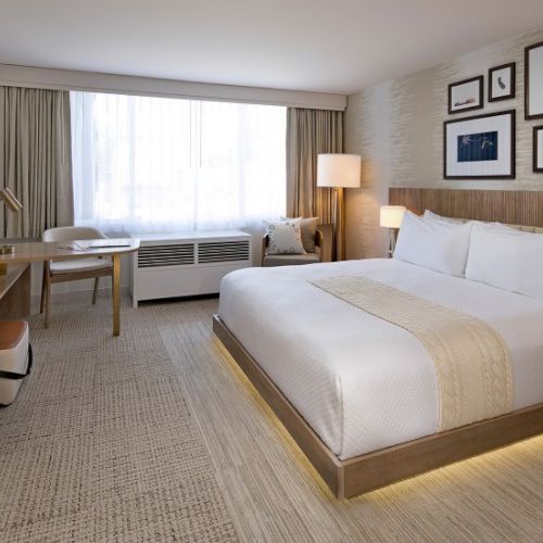 A bright hotel room with a large bed, neutral tones, a desk, seating area by the window, and framed art above the headboard end the sentence.