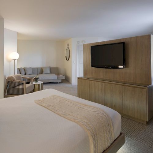 A hotel room with a large bed, a matching bench, a sitting area with a sofa, a wall-mounted TV on wood panel, and a windowed corner; calm, neutral tones.