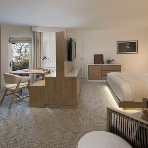 A modern hotel suite with a large bed, seating area, desk, and TV; soft lighting, neutral tones, and a balcony view, stylish and cozy.