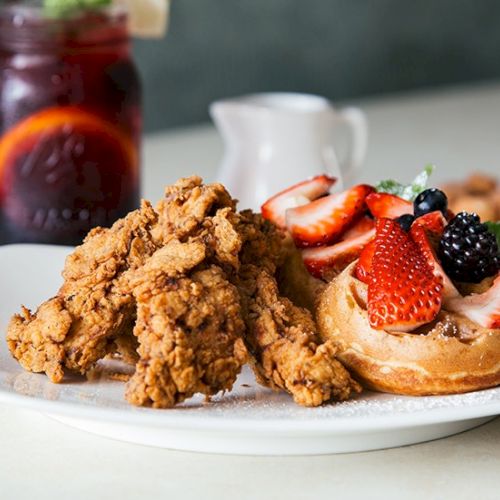 Delicious fried chicken with crispy coating served on a plate, with a side of fries and a tomato slice, a bottle of sauce nearby.