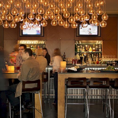 A cozy caf&eacute; scene with people chatting at a counter, warm lighting, and a chandelier; someone appears to be ordering or paying at the bar.
