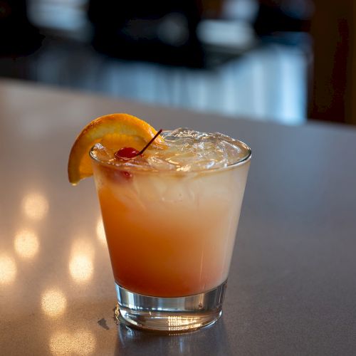 A cocktail with an orange slice, served on a wooden bar. Warm, soft lights glow in the background, creating a cozy lounge vibe.
