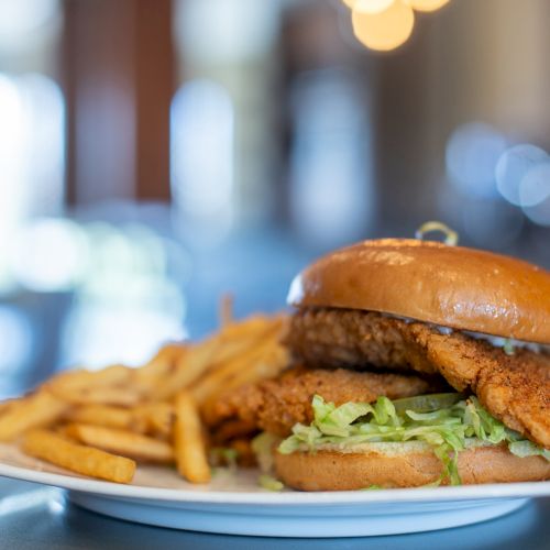 A sandwich with a crispy fried chicken patty, lettuce, and sauce sits on a plate at a cafe, with a blurred city background.