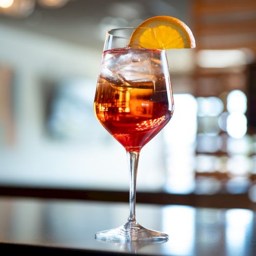 A glass of colorful cocktail with orange slice garnish sits on a bar counter, blurred bar background behind it. For 140 characters: A vibrant cocktail with orange slice on a bar, glimmering in warm light, inviting a relaxing moment.