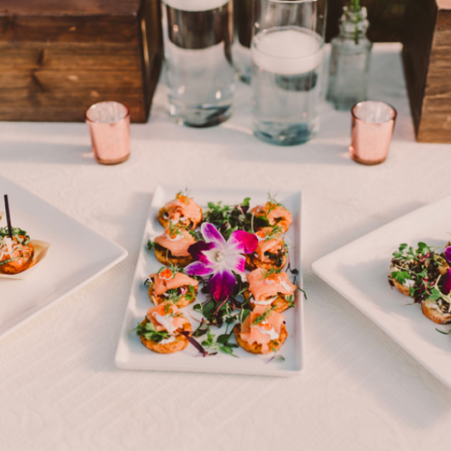 A row of small plants and candles with a central colorful flower arrangement on a white table, flanked by decorative jars and soft pastel accents.