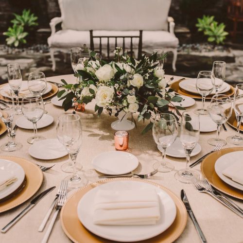 A formal dining table set with white and gold plates, glassware, napkins, and a floral centerpiece in an elegant outdoor venue.