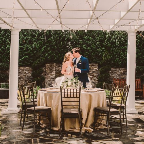 A couple stands by a round, draped table under a white trellis, kissing as they prepare to toast, overlooking a garden patio.