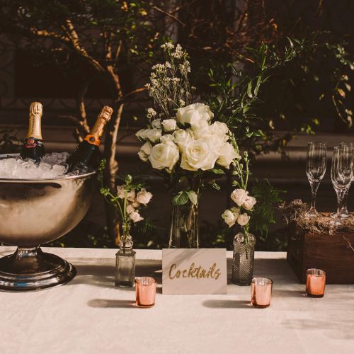 A champagne bar setup with a silver punch bowl, flowers, bottles, and small pink candles, plus a &ldquo;Cocktails&rdquo; sign on a white-covered table.