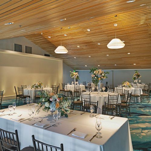 A elegantly set banquet hall with white tablecloths, floral centerpieces, wine glasses, and wooden chairs, ready for a formal event.