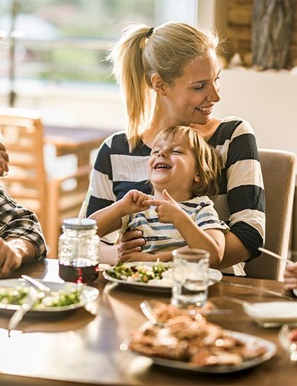 A happy family at a dining table sharing a meal, parents smiling with two kids as they enjoy food together.