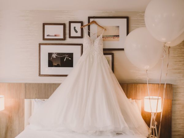 A white wedding dress hangs on a wall above a bed, framed photos, and balloons float nearby in a softly lit room.