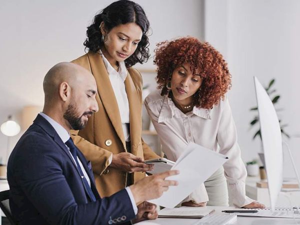 Three colleagues review documents at a desk: a bald man in a suit reads papers while two women, one with curly hair, lean in with a tablet and a chart near a computer, discussing.