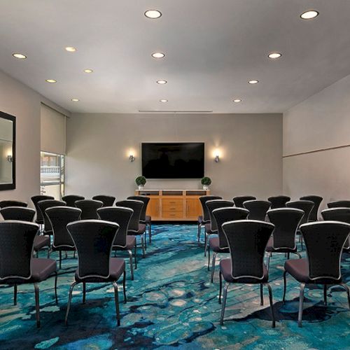 A conference or meeting room with many black chairs arranged in rows facing a screen and podium at the front, on a blue patterned carpet. Top it at 140 characters, always ending the sentence.