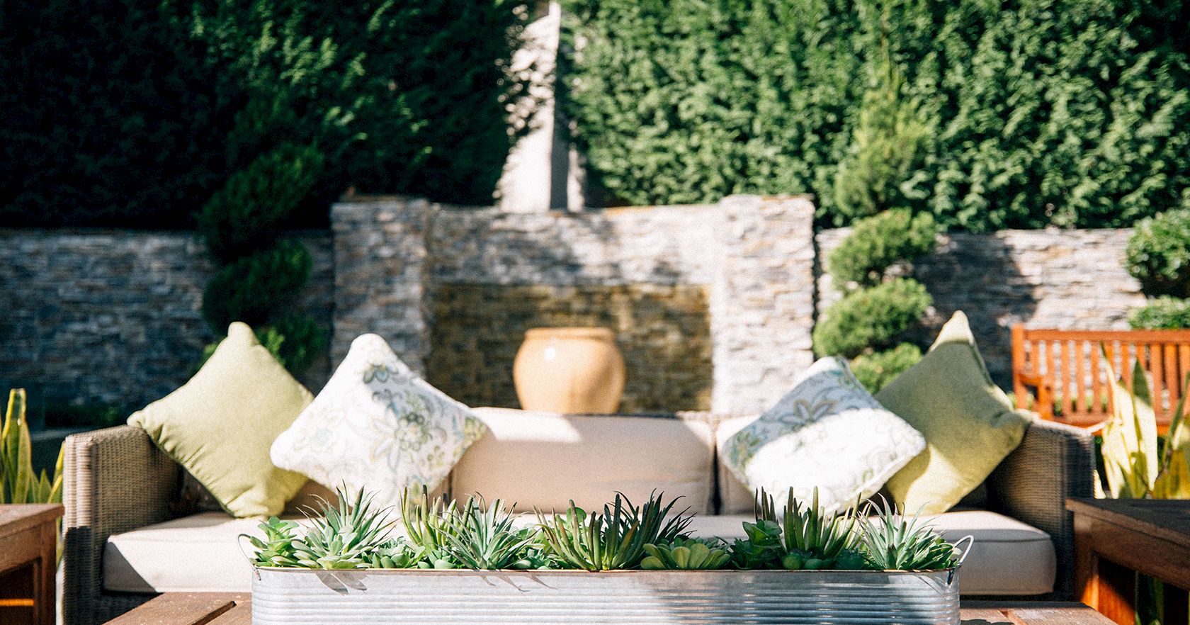 An outdoor seating area with a cushioned sofa, throw pillows, and a rectangular planter of succulents on a wooden deck, backed by stone walls and greenery.